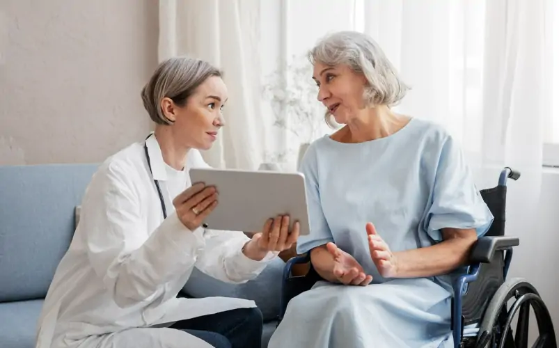 Smiling cheerful young female doctor in white uniform visiting disabled elderly adult patient at home, asking about health at check up meeting, geriatric healthcare assistance concept
